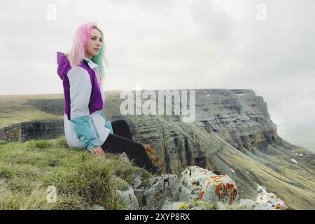 Ein Mädchen-Reisende mit bunten Haaren sitzt am Rande einer Klippe und blickt auf den Horizont auf einem Hintergrund eines felsigen Plateau Stockfoto