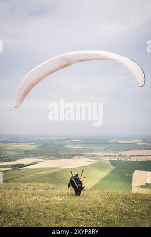 Ein Mann Gleitschirm, der vom Rand des Berges mit Feldern im Hintergrund abheben. Gleitschirmfliegen Stockfoto