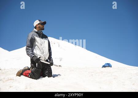 Training zum Korrigieren des Rutschens an Hanglagen oder Gletschern mit Hilfe einer Eispistole. Ein voll ausgestatteter Backpacker kniet auf einem schneebedeckten Hang in den Bergen in der Nähe Stockfoto