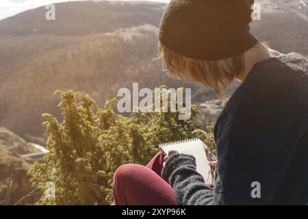 Porträt eines Hipster-Mädchens mit Sonnenbrille und Hut, das draußen auf einem Felsen in den Bergen vor blauem Himmel sitzt. Freiberuflicher Designer in Trav Stockfoto