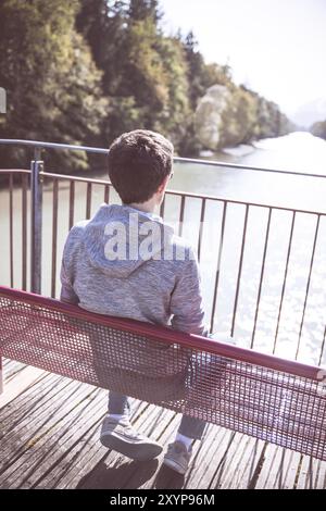 Der junge Mann sitzt auf der Bank und genießt den Blick über einen Fluss Stockfoto