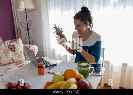 Ernährungswissenschaftler hält Ananas vor dem Laptop, während er mit frischem Obst am Tisch sitzt. Online-Beratung Stockfoto