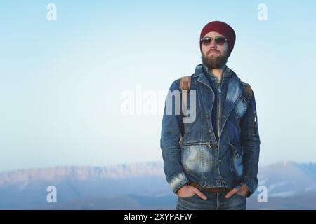 Modeporträt eines bärtigen jungen Hipster-Mannes mit Sonnenbrille, Rucksack und Hut auf einem Hintergrund mit Copyspase in den Bergen bei Sonnenuntergang, Eine Co Stockfoto