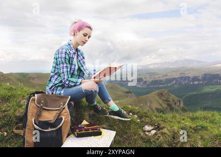 Ein Reisender sitzt in den Bergen auf dem Gras und liest ein Buch über den Hintergrund epischer Berge. Der Begriff des Lesens während der Ruhe und im Urlaub Stockfoto