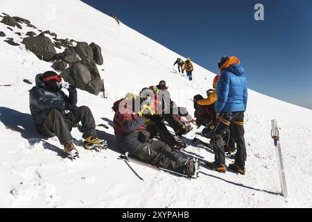 Voll ausgestattete professionelle Bergsteiger sitzen bei sonnigem Wetter auf einer verschneiten Piste. Das Konzept der kollektiven Erholung in den Bergen Stockfoto