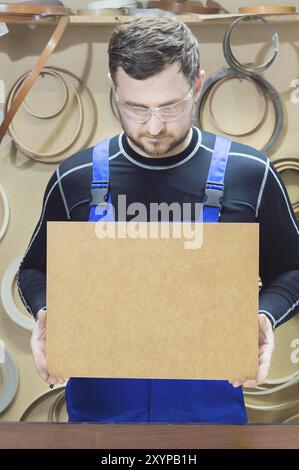 Der Hersteller von Möbeln in Spezialkleidung und Schutzbrille hält am Arbeitsplatz eine leere Platte für den Text. Handwerkliche Herstellung von Möbeln Stockfoto