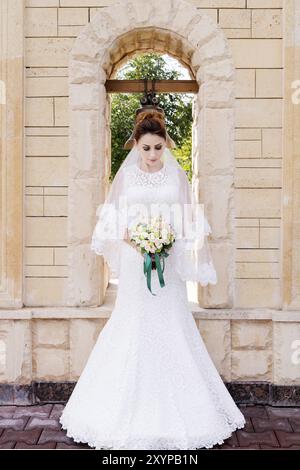 Eine schöne Braut in einem Hochzeitskleid und einem Hochzeitsstrauß in ihren Händen steht draußen vor einem Steinbogen mit einer Glocke. Das Konzept des Wedding Happi Stockfoto