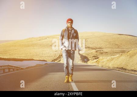 Ein stylischer bärtiger Hipster in Sonnenbrille mit einem Vintage-Rucksack spaziert an einem sonnigen Tag entlang der Asphaltstraße. Das Konzept des Anhalter- und Wanderns Stockfoto