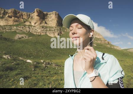 Fitness-Läuferin schließt die Augen und hört Musik über die Natur. Porträt eines schönen Mädchens mit Ohrhörern und Laufkappe. Nein Stockfoto