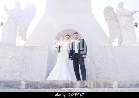 Porträt eines wunderbaren Paares, das an einem Hochzeitstag mit einem Blumenstrauß in der Hand vor dem Hintergrund eines orthodoxen christlichen Denkmals mit Engeln verbrachte. Stockfoto