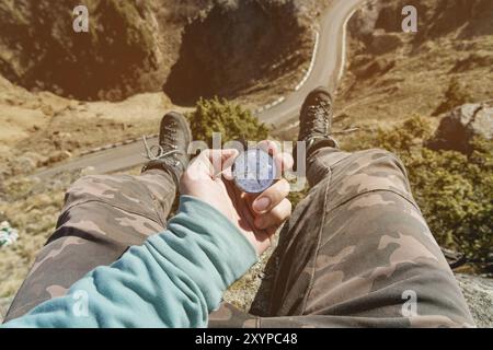 Wanderer, der auf einem hohen Felsen sitzt, hält einen Kompass vor ihren Füßen in Wanderschuhen und einer hohen Klippe mit einer Asphaltstraße darunter. Der Begriff des Recrea Stockfoto