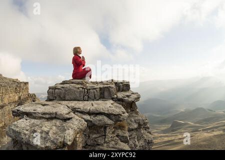 Ein schönes Mädchen meditiert in Lotusstellung, sitzt auf einem Felsen über den Wolken vor dem Hintergrund der untergehenden Sonne und des Sonnenuntergangs Stockfoto