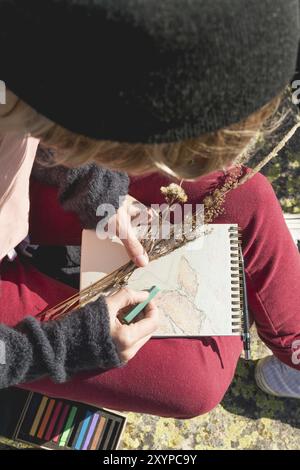 Stilvolle Mädchen Designer Maler in den Bergen sitzt auf einem Felsen zieht in einem Notizblock bei Sonnenuntergang Stockfoto