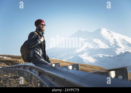 Bärtiger Touristen-Hipster-Mann in Sonnenbrille mit einem Rucksack, der auf einem Straßenrand sitzt und den Sonnenuntergang vor dem Hintergrund eines schneebedeckten M beobachtet Stockfoto