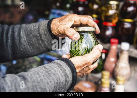 Zerknitterte alte Frauenhände halten ein Glas mit hausgemachter Marmelade aus Nadelbäumen in den Bergen Stockfoto