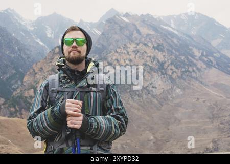 Ein Mann mit Bart und Sonnenbrille in Membran-Jacke, Hut, Rucksack und Stöcken für Nordic Walking, ein Reisender, der im offenen Ai steht Stockfoto