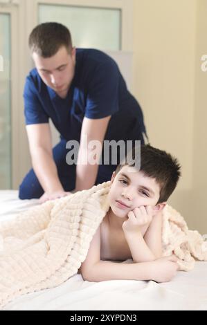 Männlicher Handtherapeut, der Kälber eines kleinen Patienten massiert. Kleiner nachdenklicher Junge beim Empfang des Massageverfahrens in der Praxis der körperlichen t Stockfoto