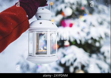 Nahaufnahme einer weiblichen Hand in einem Fäustling mit einer Hand, die eine Laterne mit Kerzen im Inneren hält, vor dem Hintergrund eines lebendigen Weihnachtsbaums in einem schneebedeckten F Stockfoto