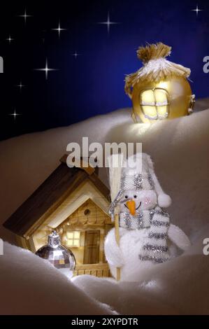 Helles konzeptionelles Weihnachtsstillleben mit einem Schneemann, der neben einem Holzhaus unter einem schneebedeckten Hügel steht. Künstlerische Weihnachtskarte, Rea Stockfoto