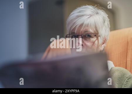 Eine Seniorin sitzt in einem Sessel und blickt über den Rand der Tageszeitung Stockfoto