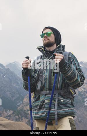 Ein Mann mit Bart und Sonnenbrille in Membran-Jacke, Hut, Rucksack und Stöcken für Nordic Walking, ein Reisender, der im offenen Ai steht Stockfoto