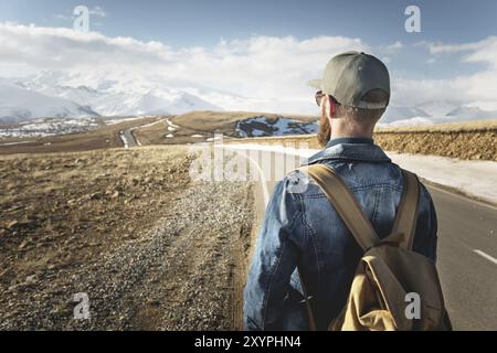 Ein bärtiger Mann in einer Mütze mit einem Rucksack, bereit, einen langen Weg zu gehen. Ein Mann auf einer Landstraße vor dem Hintergrund von Bergen und Wolken Stockfoto