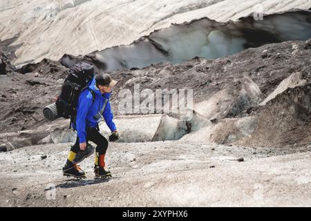 Ein Bergsteiger mit Rucksack läuft in Steigeisen entlang eines staubigen Gletschers mit Gehwegen in den Händen zwischen Rissen im Berg Stockfoto