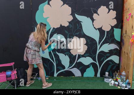 Southend on Sea, Großbritannien. 30. August 2024. Eine Frau malt Blätter auf ein großes Blumengemälde an einer schwarzen Wand, umgeben von Malvorräten und künstlichem Gras. Die Szene fängt den kreativen Prozess und die Transformation des öffentlichen Raums durch Kunst ein. City Jam Day One endet mit Künstlern, die an ihrer kreativen Arbeit arbeiten. Penelope Barritt/Alamy Live News Stockfoto