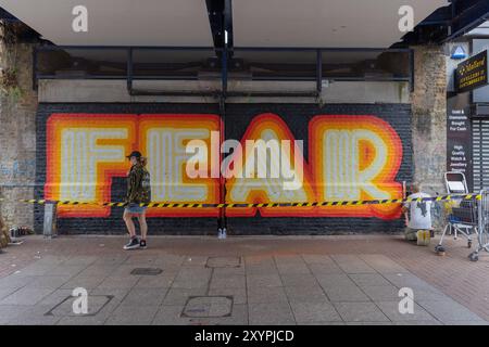 Southend on Sea, Großbritannien. 30. August 2024. Ein markantes Wandgemälde mit dem Wort „FURCHT“ in Großbuchstaben, gemalt auf einer Ziegelwand unter einem überdachten Gang. Der gestreifte Schriftzug ist rot und gelb umrandet und mit Warnband umrandet. Malmittel und eine Leiter deuten darauf hin, dass das Kunstwerk in Bearbeitung ist. Eine Person steht vor dem Wandgemälde und fügt Maßstab und Kontext hinzu. City Jam Day One endet mit Künstlern, die an ihrer kreativen Arbeit arbeiten. Penelope Barritt/Alamy Live News Stockfoto