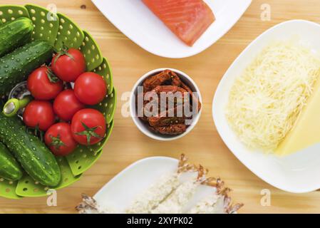 Stillleben roher Lebensmittel auf weißen Tellern auf einem Holztisch. Gefrorener Lachs auf einem Teller neben Gurken und Tomaten geriebener Käse und rohe Garnelen in bre Stockfoto