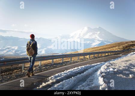 Ein stylischer bärtiger Hipster in Sonnenbrille mit einem Vintage-Rucksack spaziert an einem sonnigen Tag entlang der Asphaltstraße. Das Konzept des Anhalter- und Wanderns Stockfoto