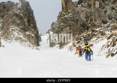 Extremskifahrer und Snowboarder klettern auf den Gipfel entlang des Couloirs zwischen den Felsen, bevor die Freeride-Abfahrt im Hinterland stattfindet. Das Konzept einer Gruppe e Stockfoto