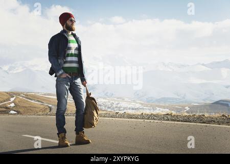 Ein stylischer bärtiger Hipster in Sonnenbrille mit einem Vintage-Rucksack steht an einem sonnigen Tag auf einem Landstraßenasphalt. Das Konzept des Anhalter- und Wanderns Stockfoto