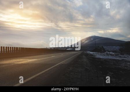 Eine Landasphaltstraße beleuchtet von den Sonnenstrahlen durch die Wolken, die zum Berg führen, auf dem die Sonnenstrahlen am Abend fallen Stockfoto