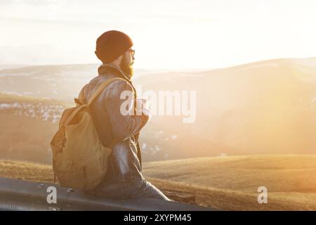 Ein bärtiger Touristen-Hipster-Mann mit Hut und Rucksack sitzt auf einer Straße und beobachtet den Sonnenuntergang vor dem Hintergrund einer Schneekappe Stockfoto