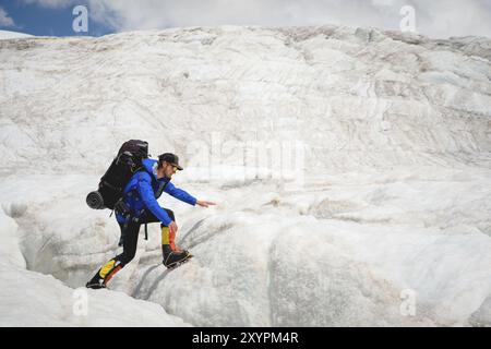 Ein Bergsteiger mit Rucksack läuft in Steigeisen entlang eines staubigen Gletschers mit Gehwegen in den Händen zwischen Rissen im Berg Stockfoto