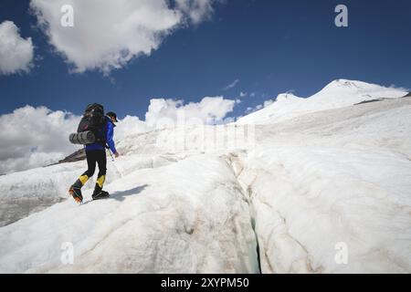 Ein Bergsteiger mit Rucksack läuft in Steigeisen entlang eines staubigen Gletschers mit Gehwegen in den Händen zwischen Rissen im Berg Stockfoto