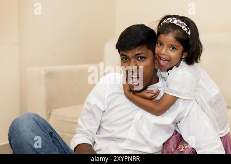 Ein Vater und seine kleine Tochter teilen einen Moment der Freude, während sie ihn spielerisch von hinten umarmt und sich gegenseitig alberne Gesichter gibt, die ein Herz erschaffen Stockfoto