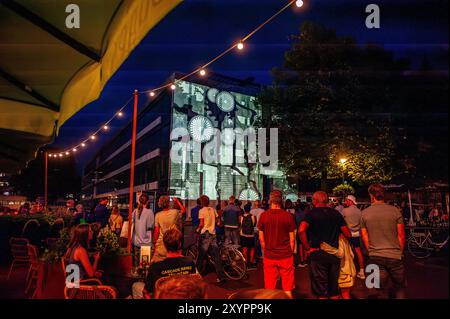 Arnhem, Gelderland, Niederlande. August 2024. Die Menschen betrachten die Bilder, die an die Wand des Gebäudes projiziert werden. An die 80. Schlacht von Arnheim erinnert man, in der Abenddämmerung erleuchten spezielle Projektionen Gebäude im Stadtzentrum. Die Schlacht bei Arnheim wurde während der Operation Market Garden ausgetragen. Die Operation Market sollte die deutschen Truppen überraschen und Arnheim und seine wichtigen Brücken über den Rhein erobern, bevor sie von den Verteidigern zerstört werden konnten. Es war der größte Luftangriff in der Geschichte und einer der ehrgeizigsten alliierten Operationen des Zweiten Weltkriegs. (Cre Stockfoto