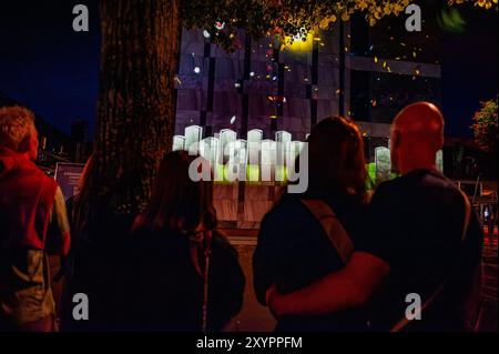 Arnhem, Gelderland, Niederlande. August 2024. Ein Paar hat gesehen, wie er sich umarmt hat und die Projektion von Bildern beobachtet hat. An die 80. Schlacht von Arnheim erinnert man, in der Abenddämmerung erleuchten spezielle Projektionen Gebäude im Stadtzentrum. Die Schlacht bei Arnheim wurde während der Operation Market Garden ausgetragen. Die Operation Market sollte die deutschen Truppen überraschen und Arnheim und seine wichtigen Brücken über den Rhein erobern, bevor sie von den Verteidigern zerstört werden konnten. Es war der größte Luftangriff in der Geschichte und einer der ehrgeizigsten alliierten Operationen des Zweiten Weltkriegs. (Guthaben Stockfoto