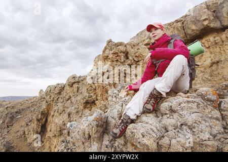 Die Reisetagerin in roter Jacke mit Rucksack und Mütze sieht auf einem Felsen hoch in den Bergen des kaukasiens auf einer untergehenden Sonne auf dem Felsen aus Stockfoto