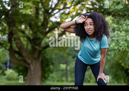 Afroamerikanerin in Sportbekleidung macht eine Pause während des Outdoor-Trainings im Park. Sie scheint entschlossen, aber ermüdet zu sein und zeigt Motivation und Ausdauer vor üppig grünem Hintergrund unter sonnigem Sommerhimmel. Stockfoto