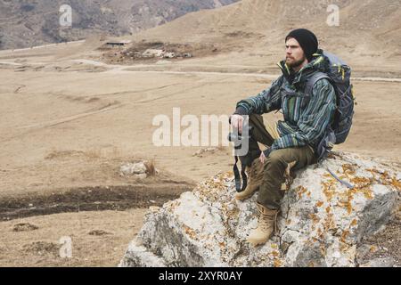 Müder bärtiger Hipster mit Fernglas in den Händen sitzt auf einem Stein zwischen den kaukasischen Bergen und blickt in die Ferne Stockfoto