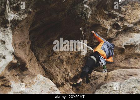 Hipster, Kletterer im Alter, in dem man einen wunderschönen Felsen ohne Versicherung und Helm hochklettert. Ein Bergsteiger in Hut und Daunenjacke mit Tasche für magnes Stockfoto