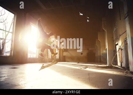 Ein junger Skater mit weißem Hut und schwarzem Sweatshirt macht einen Trick mit einem Skatesprung in einem verlassenen Gebäude im Hintergrund der untergehenden Sonne. Die Stockfoto