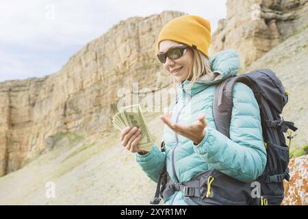 Ein Reisender mit Hut und Sonnenbrille hält hundert Dollar-Scheine in den Händen eines Ventilators vor dem Hintergrund von Felsen auf der Natur. Reisekosten Stockfoto