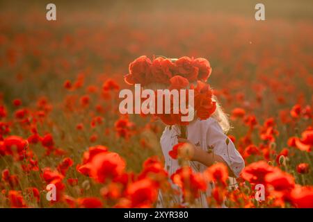 Ein junges Mädchen steht auf einem Feld roter Blumen. Sie trägt eine Blumenkrone und hält einen Strauß roter Blumen. Die Szene ist friedlich und Stockfoto