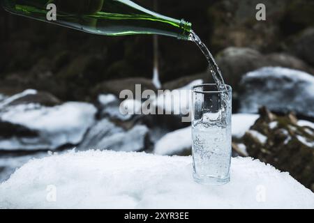 Mineralwasser wird aus einer glasgrünen Flasche in ein klares Glas-Becherglas gegossen. Ein Glas steht im Schnee. Vor dem Hintergrund eines Winters Stockfoto