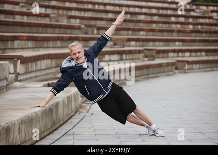 Vollständige Aufnahme eines fitten älteren Mannes in Sportbekleidung, der seitliche Plank hält, während er sich auf riesige Betonstufen des Amphitheaters lehnt, während er regelmäßig im Stadtpark, Kopierraum die Muskeln trainiert Stockfoto