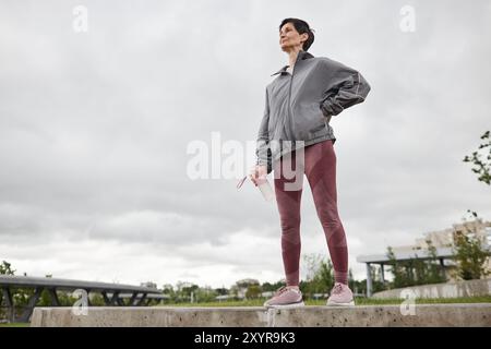 Vollständige Aufnahme einer lächelnden, fitten älteren Frau in Sportbekleidung, die eine Flasche Wasser hält, die nach dem Joggen im Stadtpark vor bedecktem Himmel ruht, Kopierraum Stockfoto